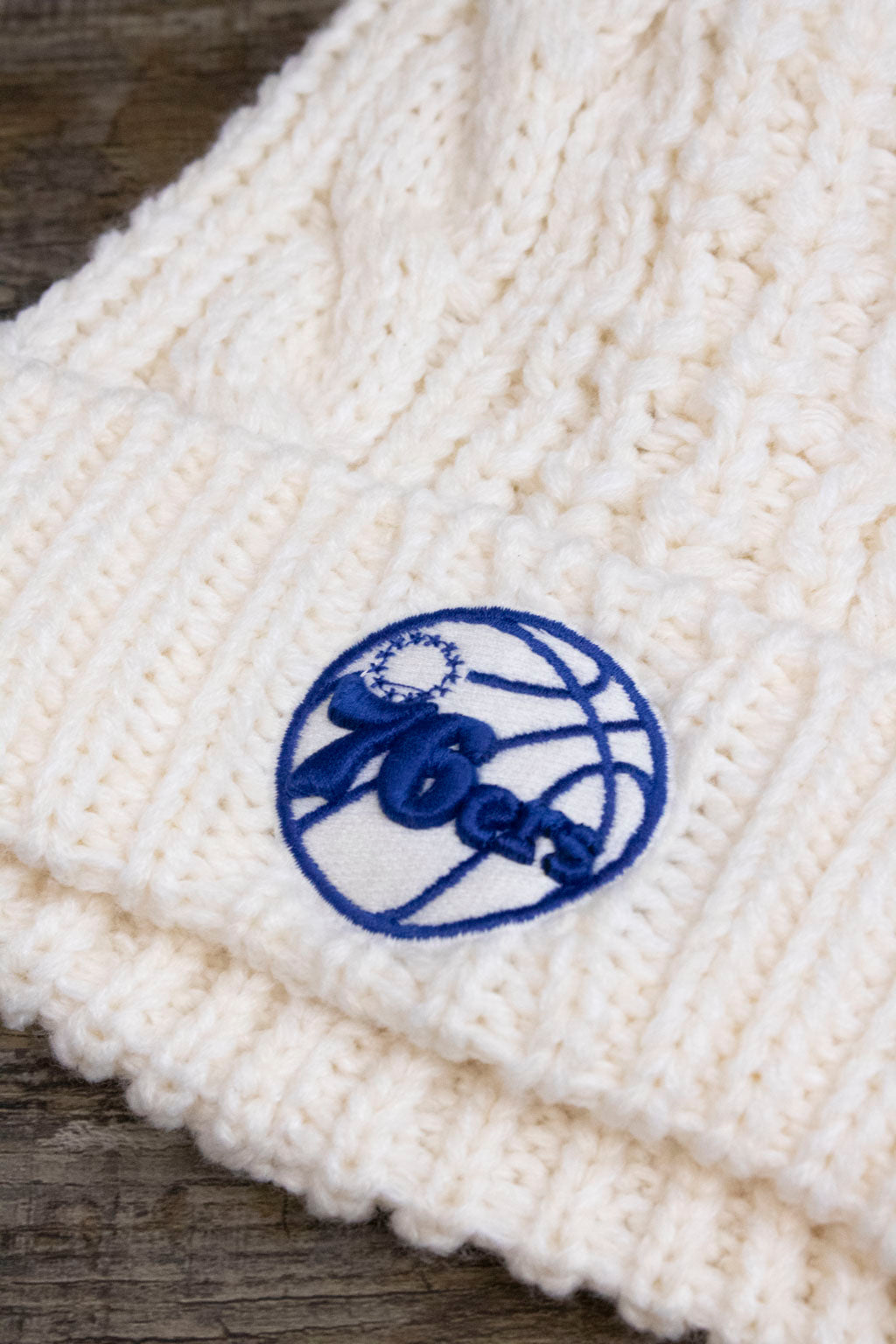 the logo on the Philadelphia 76ers Ladies Fur Pom Cream Winter Beanie | Sixers Meeko Faux Fox Fur White Pompom Beanie is made of blue and white embroidery