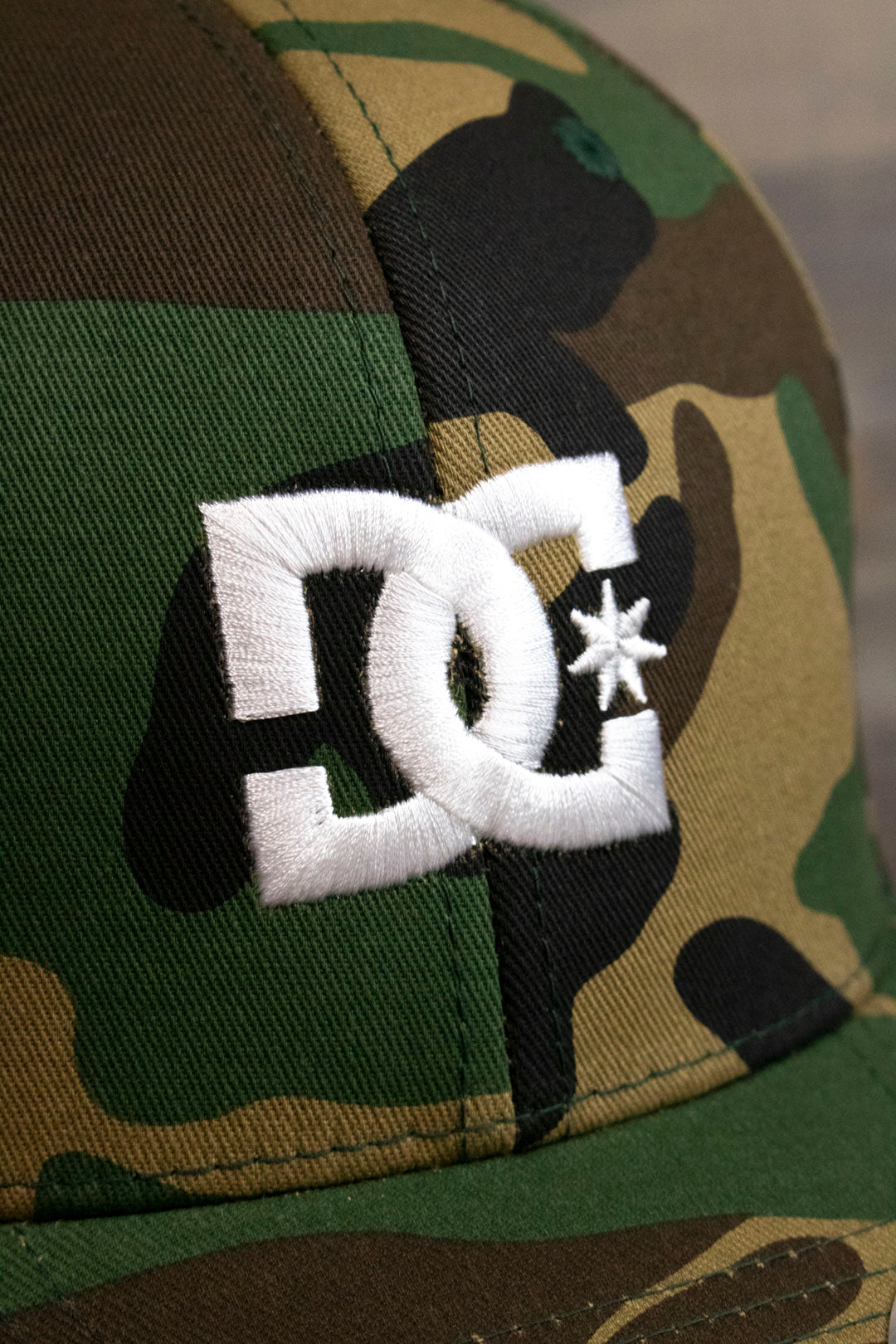 the logo on the Camouflage Print Skater Hat | DC Shoes Black Bottom Camo Flexfit Cap is made of white satin embroidery