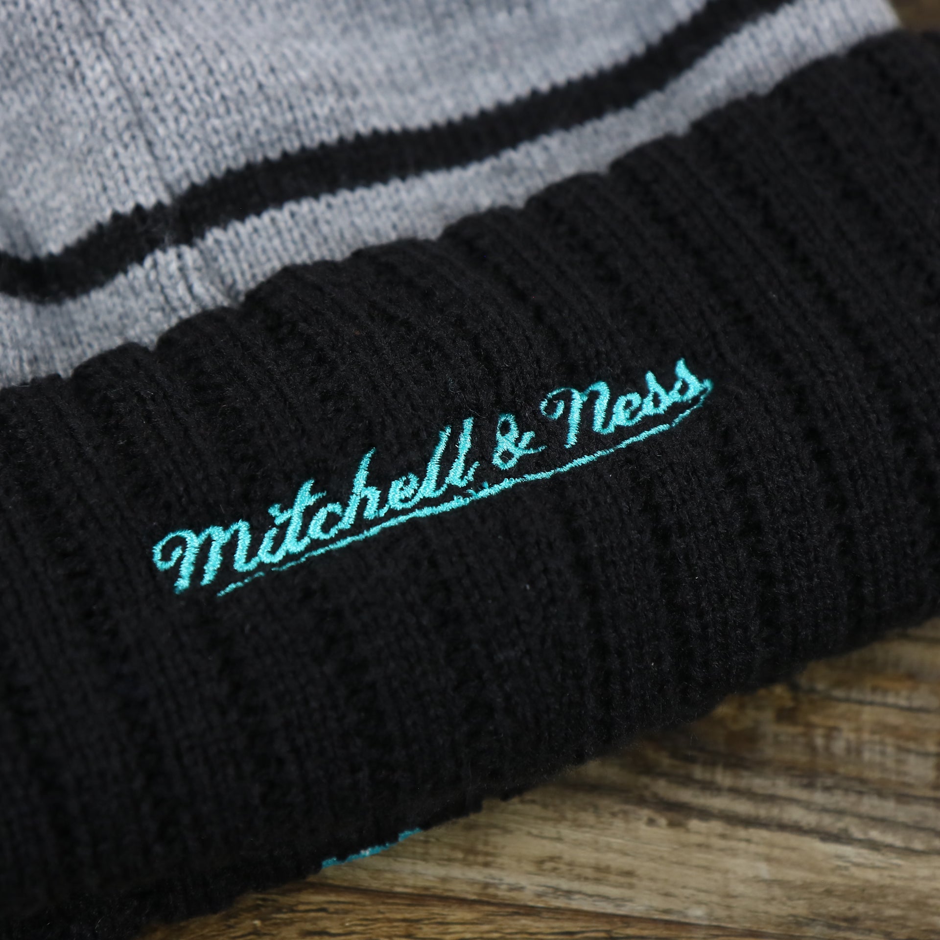 The Mitchell And Ness Script Logo on the Retro San Antonio Spurs Striped Wordmark Cuffed Pom Pom Beanie | Gray And Black Winter Beanie