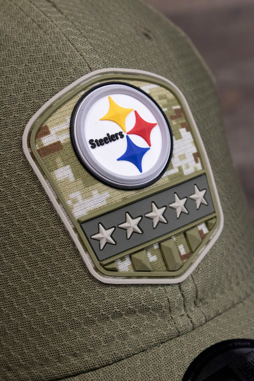 the rubberised patch on the Pittsburgh Steelers 2019 Salute To Service Dad Hat | Steelers On Field Olive Green Military Inspired Baseball Cap has stars and stripes and a round Steelers emblem on it