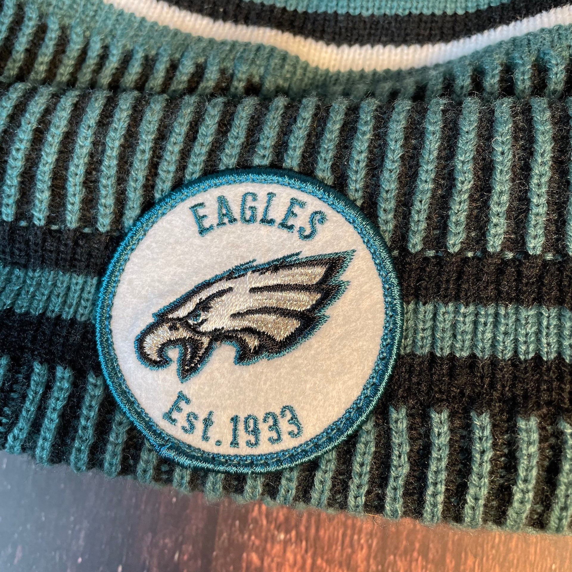 Close up on the Philadelphia Eagles On Field Eagles Colorway Striped Pom Pom Beanie front Eagles logo