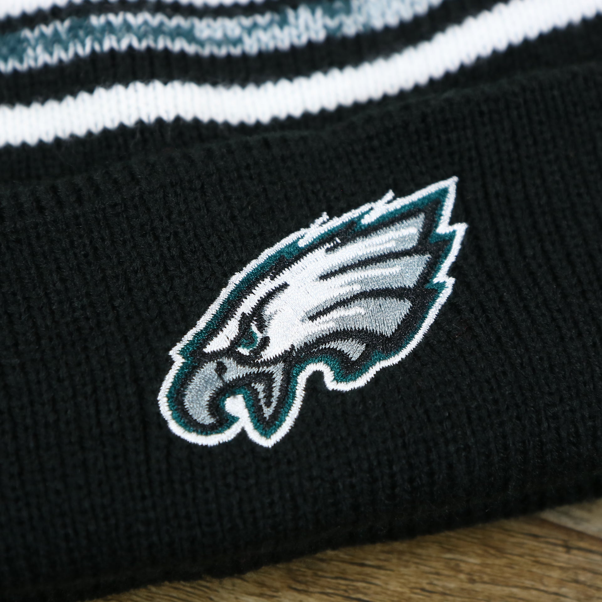 The Eagles Logo on the Kid’s Philadelphia Eagles Striped Pom Pom Winter Beanie | Black, Midnight Green, And White Beanie