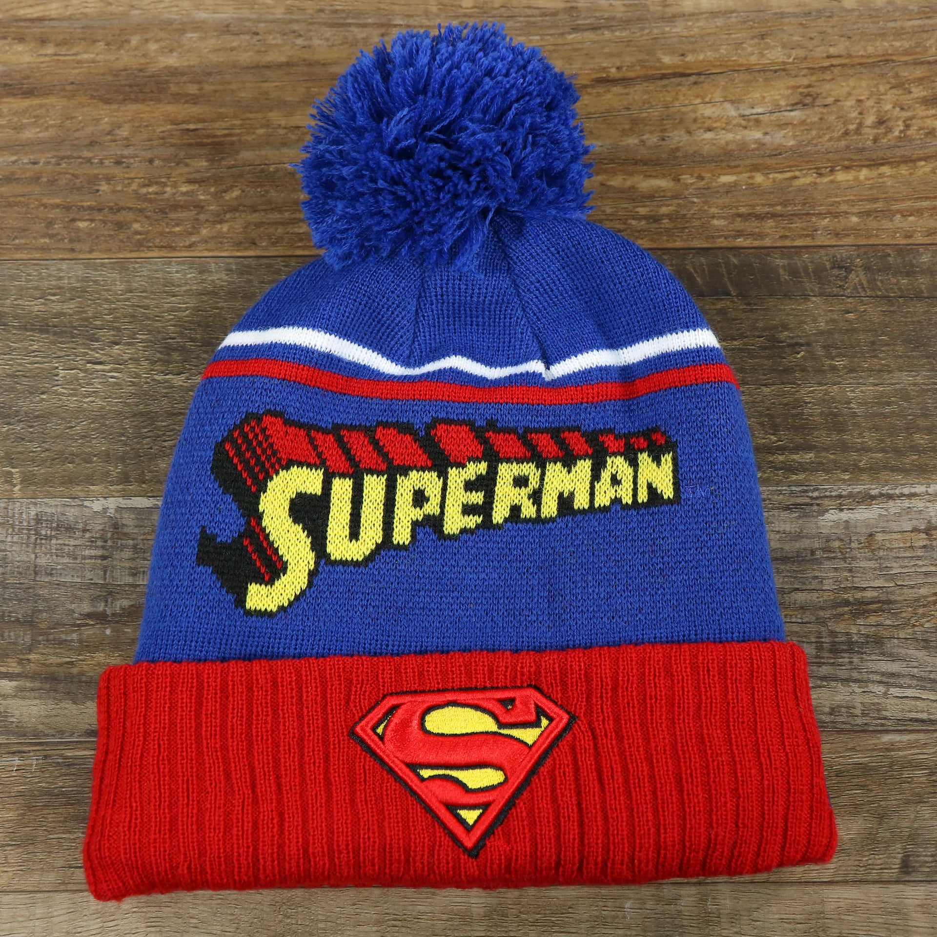 The front of the DC Comics Superman S-Shield Logo Superman Wordmark Striped Beanie With Blue Pom Pom | Blue And Red Winter Beanie