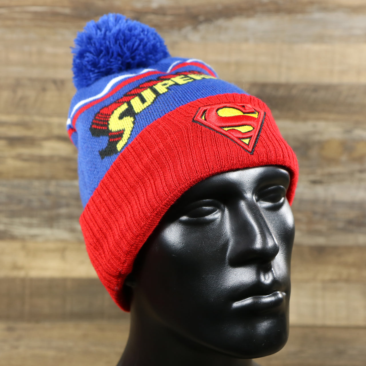 The DC Comics Superman S-Shield Logo Superman Wordmark Striped Beanie With Blue Pom Pom | Blue And Red Winter Beanie on a doll