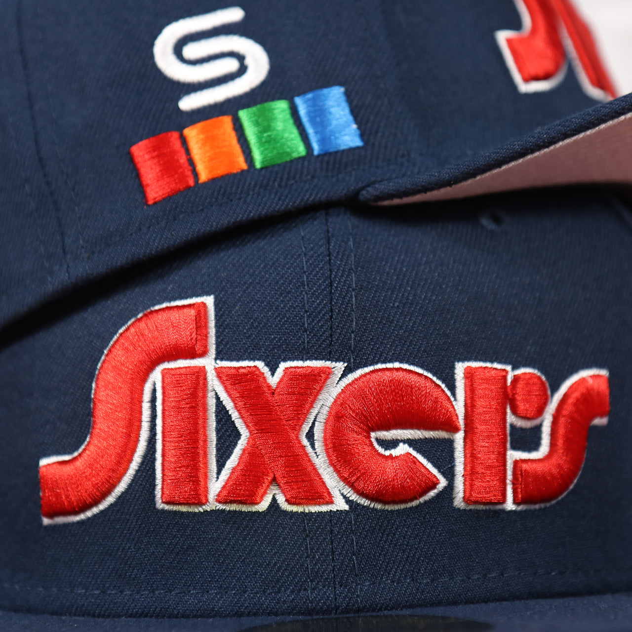 Close up of the side patch and front wordmark of the Philadelphia 76ers 2022 City Edition Sixers Script Spectrum Side Patch Navy 59Fifty Fitted Cap