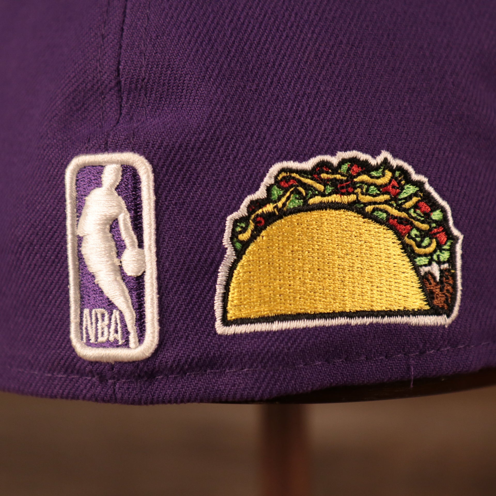 A close up shot of the NBA logo and the taco patch on the backside of the purple Los Angeles Lakers gray bottom brim all over patch embroidered 59fifty fitted hat by New Era.