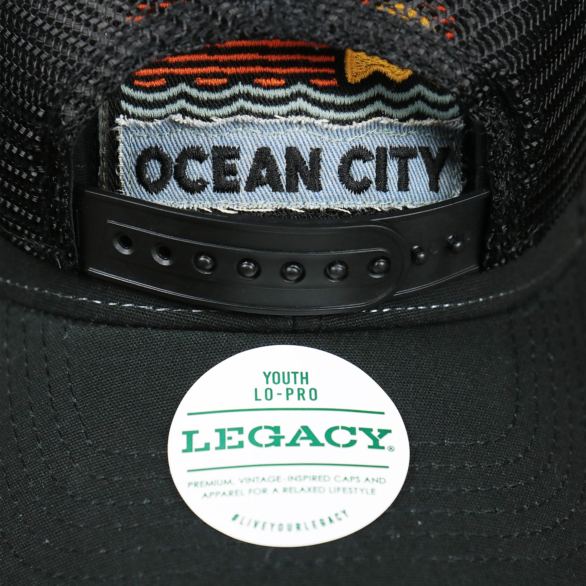 The Legacy Sticker on the Youth New Jersey Ocean City Sunset Mesh Back Trucker Hat | Black And Black Mesh Youth Trucker Hat