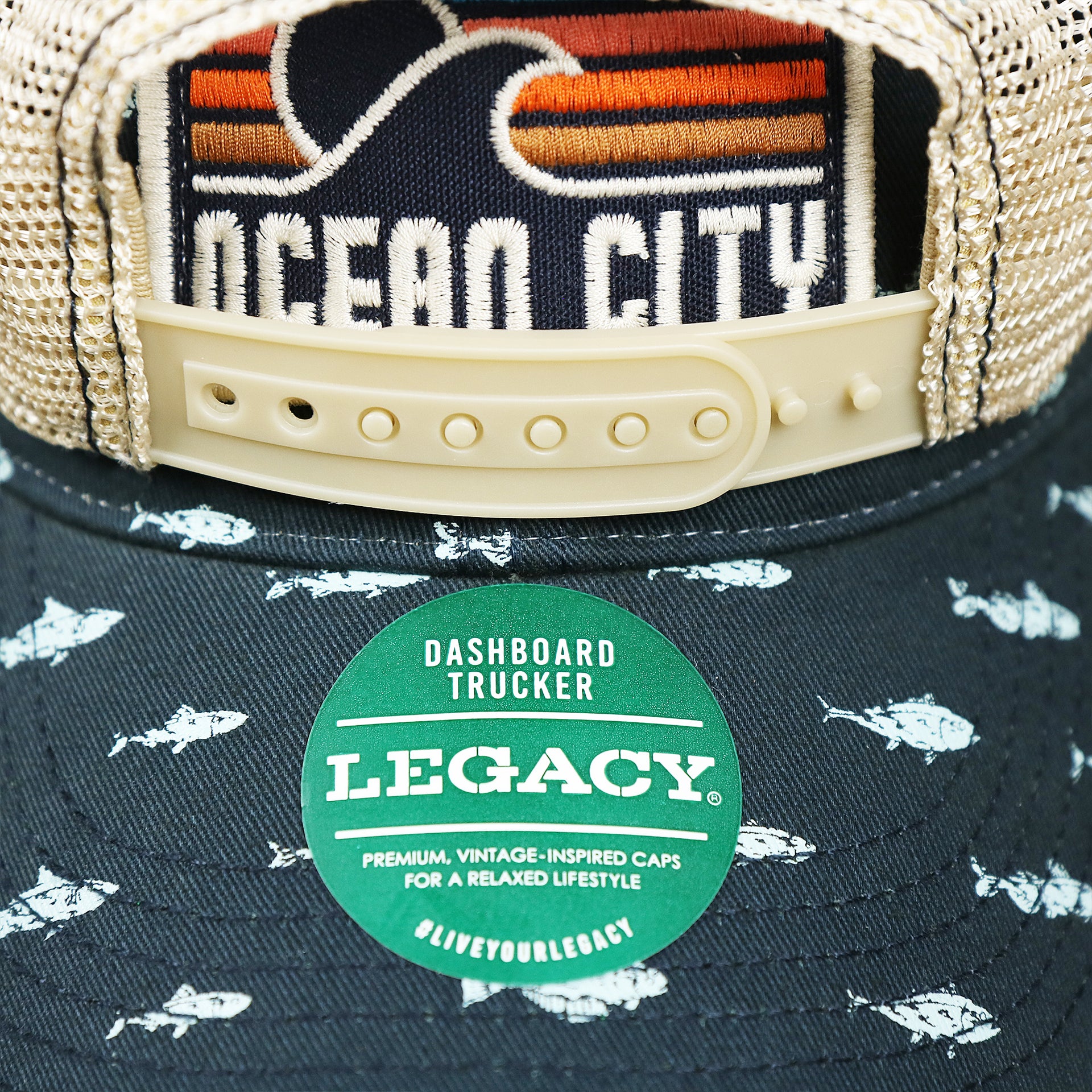 The Legacy Sticker on the Ocean City Sunset Patch Fish Print Khaki Mesh Back Trucker Hat | Navy Blue Trucker Hat