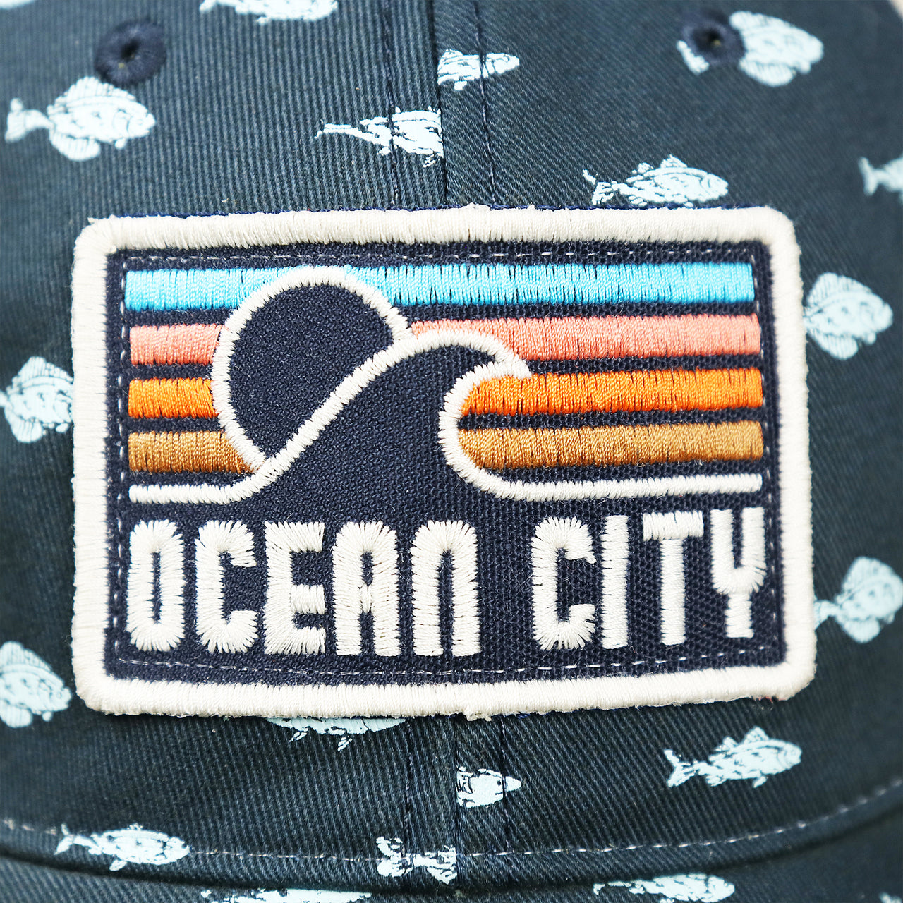 The Ocean City Sunset Logo on the Ocean City Sunset Patch Fish Print Khaki Mesh Back Trucker Hat | Navy Blue Trucker Hat